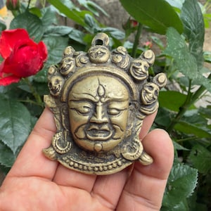 May include: A brass-colored metal pendant featuring a central face with closed eyes, a third eye on the forehead, and a crown-like arrangement of smaller faces around the perimeter. The piece has a small loop at the top for hanging or attachment.