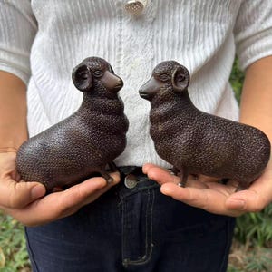 Könnte beinhalten: Zwei dunkelbraune, texturierte Schaf-Figuren mit detaillierten, geschwungenen Hörnern und wolligem Fell. Jedes Schaf steht auf vier kleinen Beinen und ist einander zugewandt. Die Figuren haben glatte Gesichter und Ohren mit matter Oberfläche.