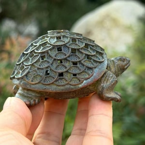 May include: A small, dark bronze-toned turtle figurine with a shell crafted from a repeating pattern of circular coin-like motifs. The surface has a weathered, textured patina with visible green oxidation, and the head and limbs are sculpted.