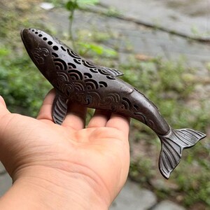 May include: A dark brown metal whale figurine featuring intricate cut-out patterns along its body. The surface is decorated with repeating wave-like motifs and small circular perforations, with detailed textures on the fins and tail fluke.