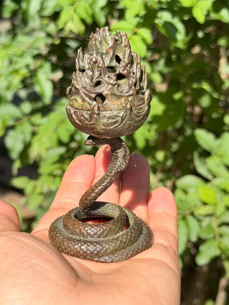 May include: A bronze-toned metal incense burner featuring a coiled snake base that supports a textured, flame-shaped upper vessel with openwork detailing. The object has a dark, weathered patina and a detailed scale pattern on the snake's body.