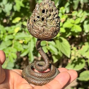 May include: A bronze-toned metal incense burner featuring a coiled snake base that supports a textured, flame-shaped top with small circular cutouts. The object has a dark, weathered patina and a detailed, scaly surface texture throughout the design.
