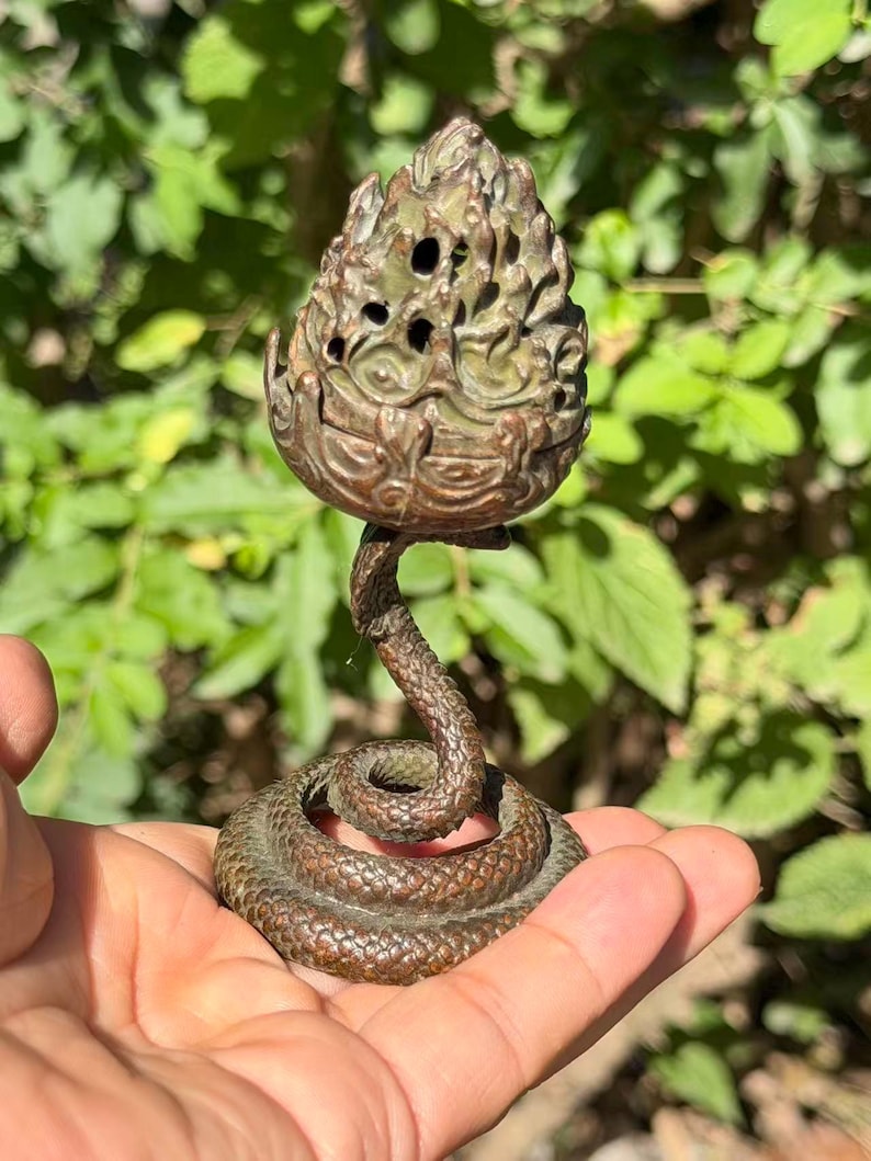 May include: A bronze-toned metal incense burner featuring a coiled snake base that supports a textured, flame-shaped top with small circular cutouts. The object has a dark, weathered patina and a detailed, scaly surface texture throughout the design.
