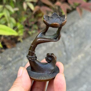 May include: A dark bronze-toned metal sculpture featuring a stylized hand extending upward from a circular base. The hand is cupped to support a lotus flower-shaped bowl at the top. The base has a radial line pattern and a small decorative element.