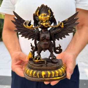 May include: A bronze-colored statue of a mythical creature with large wings, a bird-like face, and a snake held in its beak. The figure stands on a circular lotus-shaped base with gold-toned highlights, featuring intricate detailing and a dark finish.