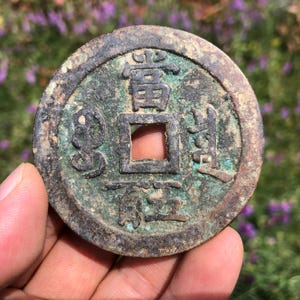 May include: Antique bronze coin with square hole in the center and Chinese characters. The coin has a weathered appearance with green and brown patina. The coin is held in a hand against a blurred background of purple flowers.