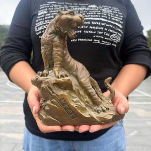 Puede incluir: Una figura de tigre color bronce sobre una base rocosa. El tigre está representado en una pose rugiente con la boca abierta, mostrando rayas y garras detalladas. Una placa rectangular en la parte frontal de la roca presenta caracteres chinos grabados.