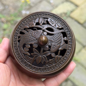 May include: Bronze-colored, round incense burner with a detailed design of two koi fish facing each other, surrounded by lotus flowers and decorative patterns. The lid features a small, round handle. The overall aesthetic is ornate and traditional.