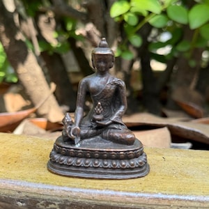 May include: A small, bronze-colored Buddha statue in a seated position. The figure is detailed with a serene expression, wearing draped robes, and holding a small object. The statue sits on a decorative base with a detailed pattern.