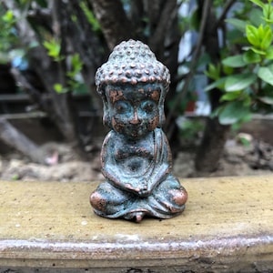 May include: A small, weathered bronze Buddha statue in a seated meditation pose. The statue has a detailed head with a textured bun and is colored with shades of green and copper. The figure is sitting on a light brown surface, with a blurred green background.