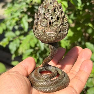 May include: A bronze-toned metal incense burner featuring a coiled snake base that rises to support a textured, openwork pinecone-shaped lid. The surface of the object displays a weathered, dark patina with intricate scale details on the snake.