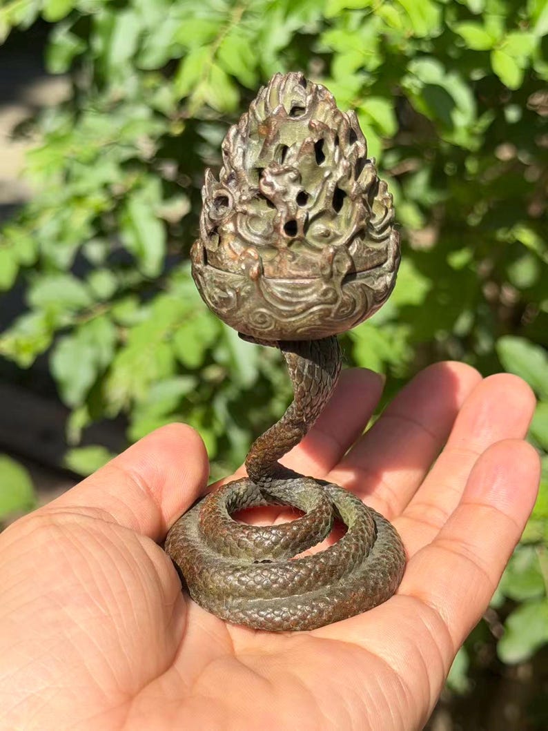 May include: A bronze-toned metal incense burner featuring a coiled snake base that rises to support a textured, openwork pinecone-shaped lid. The surface of the object displays a weathered, dark patina with intricate scale details on the snake.