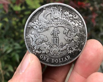 May include: A silver coin featuring a detailed dragon design, clouds, and Chinese characters. The coin is inscribed with "ONE DOLLAR" around the edge. The coin is held in a hand.