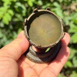 May include: A bronze-toned metal egg cup or small holder featuring a circular bowl with a scalloped rim. The base of the object is formed by a coiled snake with a textured, scale-like surface, providing a stable foundation for the cup above.
