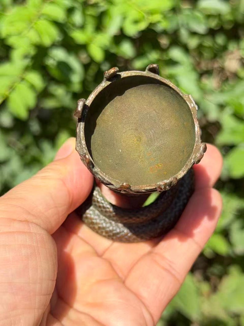 May include: A bronze-toned metal egg cup or small holder featuring a circular bowl with a scalloped rim. The base of the object is formed by a coiled snake with a textured, scale-like surface, providing a stable foundation for the cup above.