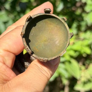 May include: A circular, metallic object with a weathered, bronze-toned finish. The item features a recessed, bowl-like center surrounded by a raised, scalloped rim with small, evenly spaced protrusions along the outer edge of the circular frame.