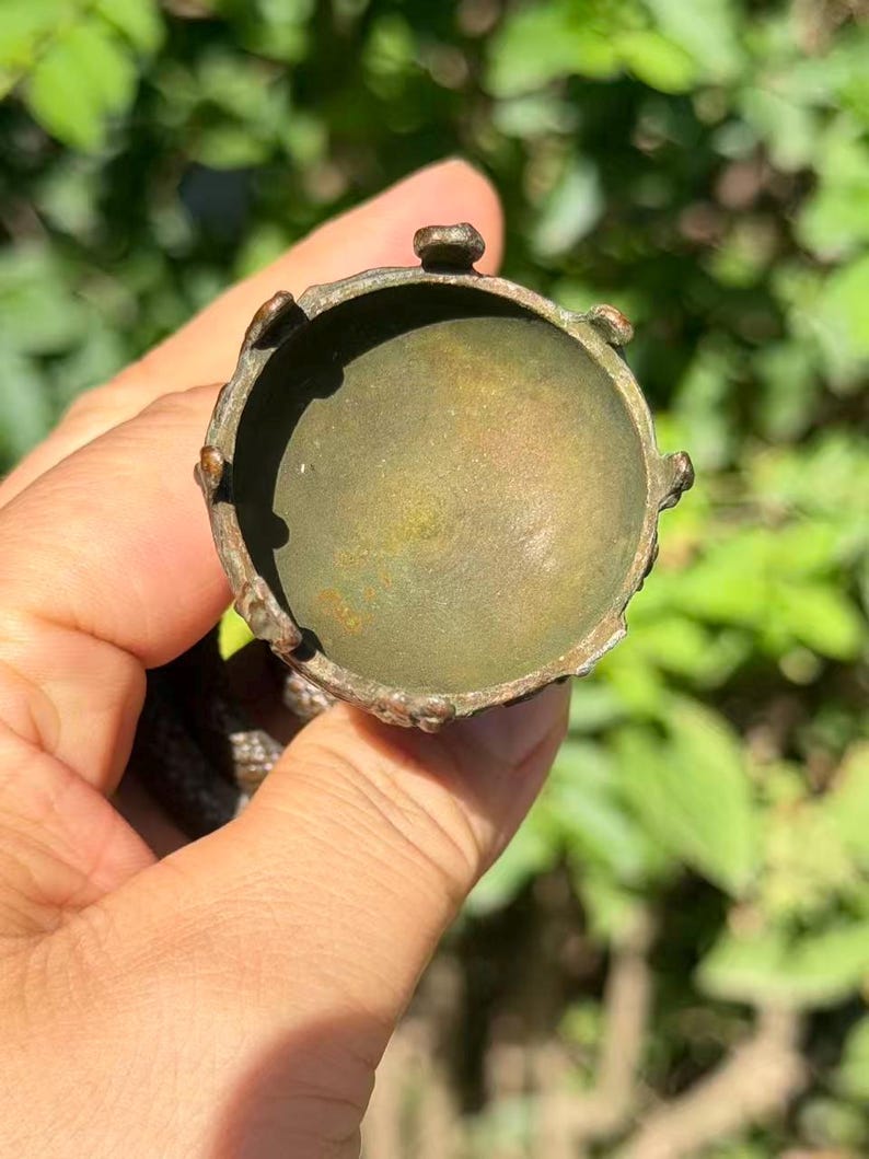 May include: A circular, metallic object with a weathered, bronze-toned finish. The item features a recessed, bowl-like center surrounded by a raised, scalloped rim with small, evenly spaced protrusions along the outer edge of the circular frame.