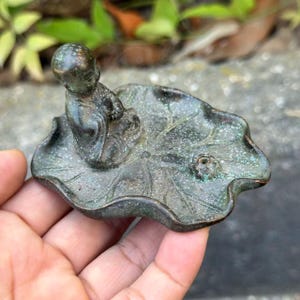 May include: A dark bronze-toned metal incense holder featuring a small, seated figure of a monk resting on a textured lotus leaf base. The surface has a weathered, speckled patina with visible veins on the leaf and a small circular opening.