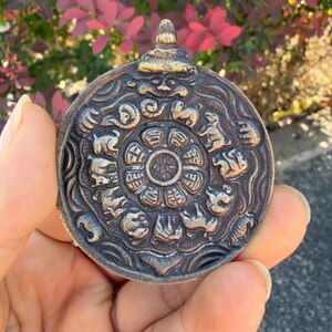 May include: A circular bronze-toned metal pendant featuring a central eight-petaled lotus design surrounded by a ring of twelve embossed animal figures. The piece has a dark, weathered patina with a small, raised, gold-toned bail at the top.