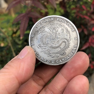 May include: A silver coin with a detailed dragon design at the centre, inscribed with "YUN-NAN PROVINCE" and "1 MACE AND 2 CANDAREENS". The coin is held between fingers, with a blurred background of green and red foliage.