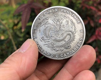 May include: A circular silver-toned coin featuring an embossed dragon at the center. The text reads YUN-NAN PROVINCE along the top edge and 7 MACE AND 2 CANDAREENS along the bottom edge. The coin has a metallic finish with visible surface details.