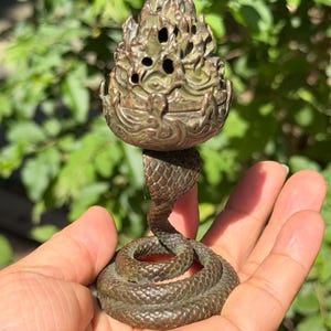 May include: A bronze-toned metal incense burner featuring a coiled snake base with textured scales supporting a decorative, perforated top shaped like a stylized flame or pinecone. The object has a dark, weathered patina finish throughout.