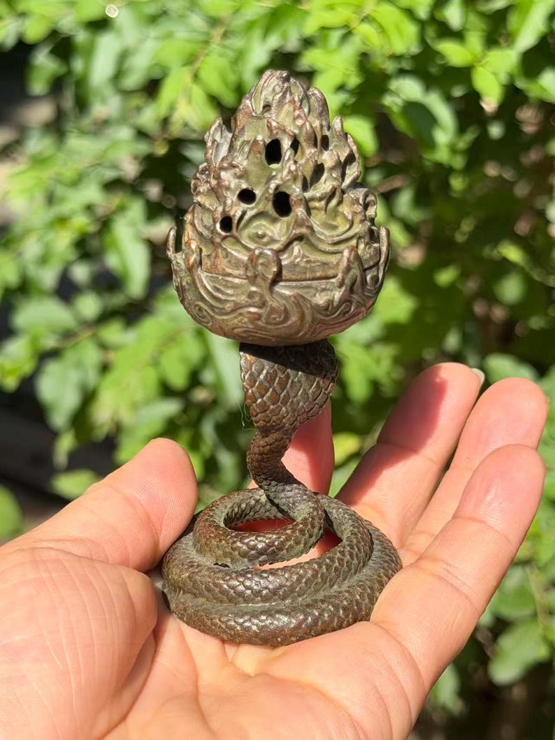 May include: A bronze-toned metal incense burner featuring a coiled snake base with textured scales supporting a decorative, perforated top shaped like a stylized flame or pinecone. The object has a dark, weathered patina finish throughout.