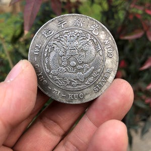 Puede incluir: Una moneda de plata con un diseño detallado de dragón en el centro, rodeada de caracteres chinos y las palabras "SILVER COIN". La moneda se sostiene en una mano, mostrando sus intrincados detalles y su apariencia envejecida.