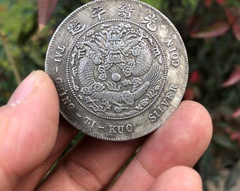 May include: A silver coin featuring a detailed dragon design at the center, surrounded by Chinese characters and the words "SILVER COIN". The coin is held in a hand, showcasing its intricate details and aged appearance.