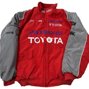 May include: Red and grey racing jacket featuring the Toyota logo and Panasonic branding. The jacket has a zip-up front, a stand-up collar, and grey sleeves. The word "DENSO" is on the collar.