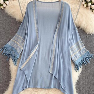 May include: A light blue open-front kimono-style top with long sleeves. The sleeves and edges are adorned with white embroidery and blue fringe. The garment is hanging on a wooden hanger.
