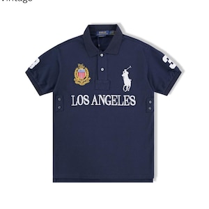 May include: A navy blue short-sleeve polo shirt featuring the text LOS ANGELES in white, a white embroidered polo player logo, a colorful crest patch, and the number 3 on each sleeve. The shirt has a buttoned collar and side vents with metal eyelets.