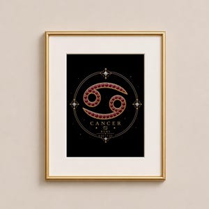Puede incluir: Impresión artística enmarcada que presenta el símbolo zodiacal de Cáncer en oro y rojo rubí sobre un fondo negro. La impresión incluye la palabra "Cáncer" y las fechas 6.22-7.22. La obra de arte está dentro de un marco dorado.