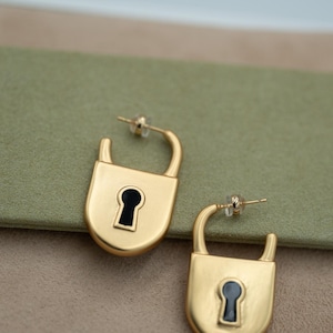 Puede incluir: Un par de pendientes dorados con forma de candado con detalles de ojo de cerradura negros. Los pendientes se exhiben sobre una superficie verde y beige, mostrando su diseño y color.