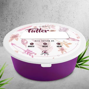 Puede incluir: Un recipiente redondo morado para comida de mascotas con tapa blanca. La tapa presenta diseños florales y la palabra alemana "Futter" con texto que indica las horas de alimentación: mañana, mediodía y tarde. El recipiente tiene un área de etiqueta blanca.