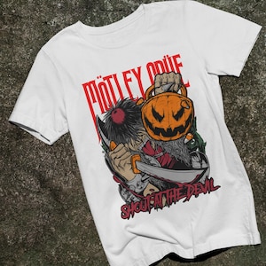 以下が含まれることがあります： ハロウィンのカボチャと大きなナイフを持つキャラクターのグラフィックプリントが施された白い半袖Tシャツ。グラフィックの上部には赤色で「Mötley Crüe」、下部には赤色で「Shout at the Devil」というテキストがプリントされています。