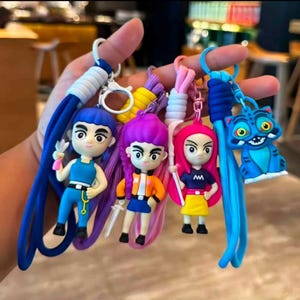May include: A selection of vibrant keyrings showcasing cartoon characters and a blue cat. The keyrings display diverse hair colours, outfits, and accessories. Each keyring includes a long, braided cord in coordinating colours.