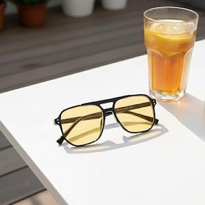 May include: Black-framed sunglasses with yellow lenses sit on a white surface beside a glass of iced tea with a lemon slice. The sunglasses have a square shape and a double bridge. The scene is lit by natural sunlight.