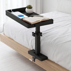 May include: A black adjustable bedside tray table with a clamp mechanism for attachment to a bed frame. The rectangular surface holds a white potted succulent, a smartphone, a silver pen, and an open book with cream-colored pages.
