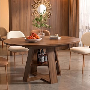 May include: A round wooden dining table with a central pedestal base featuring an integrated storage shelf. The table surface holds a white bowl filled with red apples and a glass vase with green foliage. Three chairs with cream textured upholstery surround it.