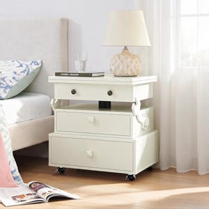 May include: A white three-tiered bedside table with a top drawer featuring two dark round knobs, a middle drawer with a square knob, and a bottom drawer with a square knob. The unit is supported by a central black column and four small black wheels.