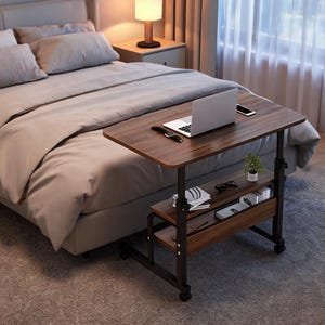 May include: A mobile overbed table with a dark wood grain finish top and two lower storage shelves. The frame is constructed from black metal with four caster wheels. The table holds a silver laptop, a smartphone, and a small potted plant.