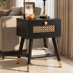 May include: A black nightstand featuring a single drawer with a gold-toned geometric lattice front panel and a small gold knob. The piece stands on four tapered black legs with gold-finished feet and includes a lower shelf for storage.