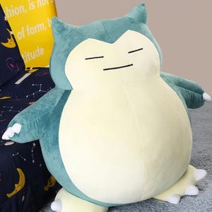 May include: A large plush toy in the shape of a Snorlax character, featuring a light blue and cream color scheme. The toy has closed eyes and small arms and feet. The background includes a yellow pillow with text.