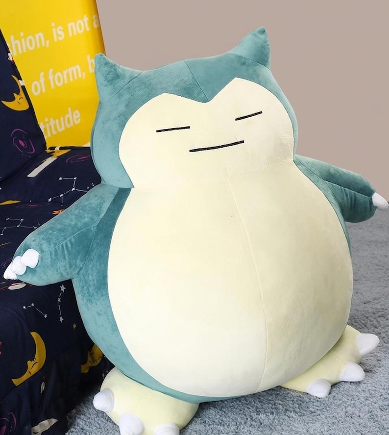 May include: A large plush toy in the shape of a Snorlax character, featuring a light blue and cream color scheme. The toy has closed eyes and small arms and feet. The background includes a yellow pillow with text.
