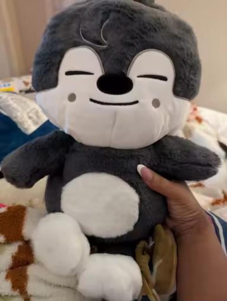 Puede incluir: Un peluche con un personaje de dibujos animados con cuerpo gris, cara blanca y detalles blancos. El juguete tiene los ojos cerrados, una nariz negra y una expresi&oacute;n sonriente. El peluche es suave y tierno.