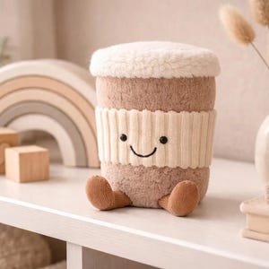 May include: A soft toy coffee cup with a smiling face, a cream-coloured knitted band, and brown feet. The cup has a fluffy white top and sits on a white shelf. Other items on the shelf include wooden blocks, a rainbow arch, and a vase.
