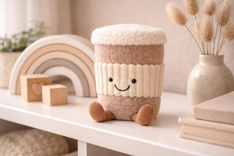 May include: A soft toy coffee cup with a smiling face, a cream-coloured knitted band, and brown feet. The cup has a fluffy white top and sits on a white shelf. Other items on the shelf include wooden blocks, a rainbow arch, and a vase.