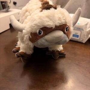 May include: A plush toy resembling a bison with large, curved horns. The toy features a cream-colored body with brown accents on the head, back, and feet. It has a gray snout and large, dark eyes, giving it a friendly appearance.