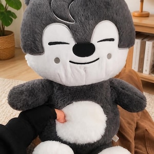 Puede incluir: Un peluche de lobo gris con detalles blancos y una cara sonriente. El peluche tiene una barriga blanca y patas blancas. El juguete est&aacute; siendo sostenido por una persona con una manga negra.